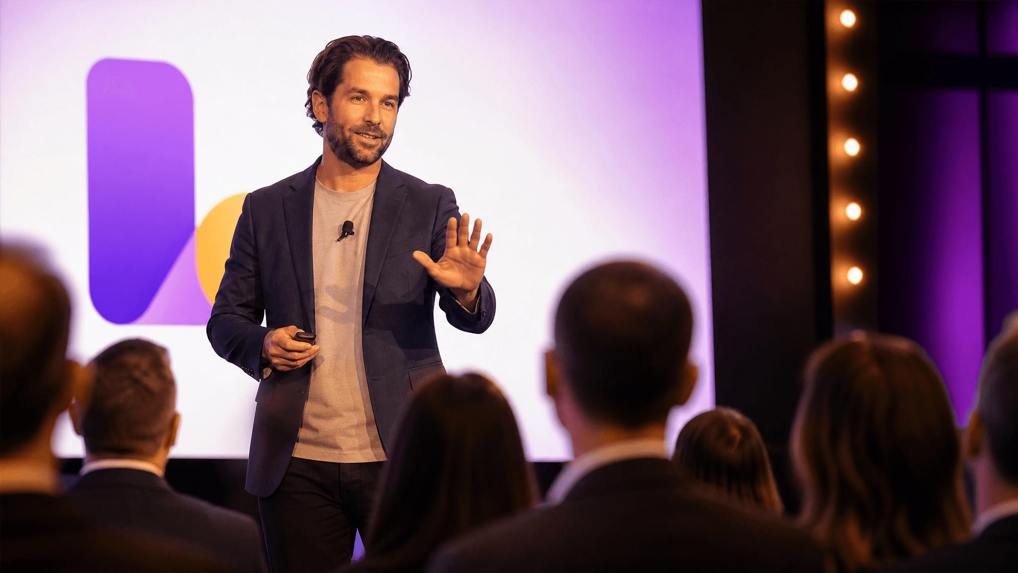 A conference speaker on stage raising a hand in a calm slow-down gesture to a blurred dental audience